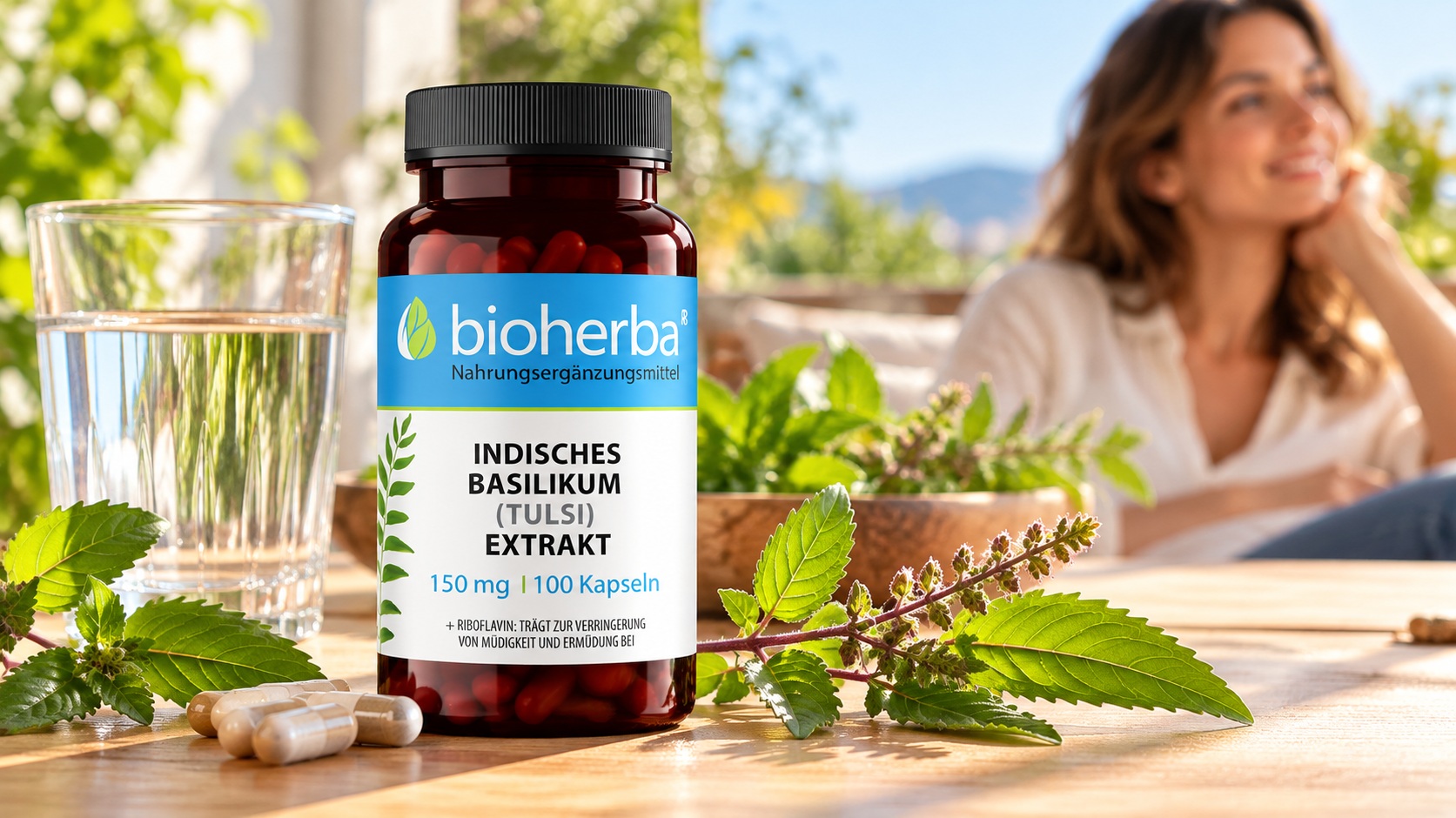 Bioherba Indisches Basilikum (Tulsi) Extrakt 150 mg &ndash; 100 Kapseln auf einem Holztisch neben Wasserglas und Tulsi-Zweigen, mit einer Frau im Hintergrund.